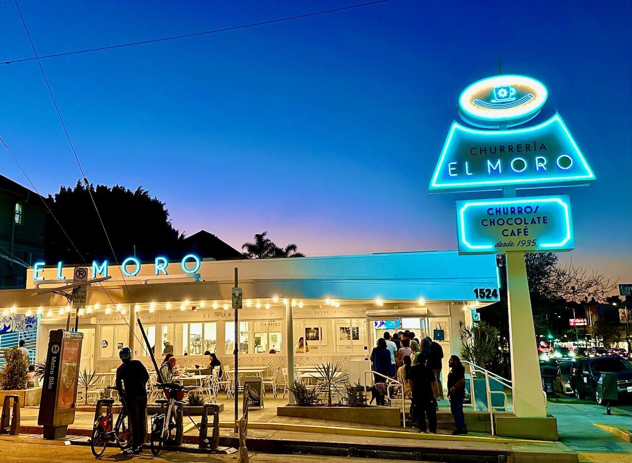 Churrería El Moro's aqua neon sign glowing at dusk above its striped-awning storefront on Sunset Boulevard, with diners at outdoor tables and bikes parked at the curb