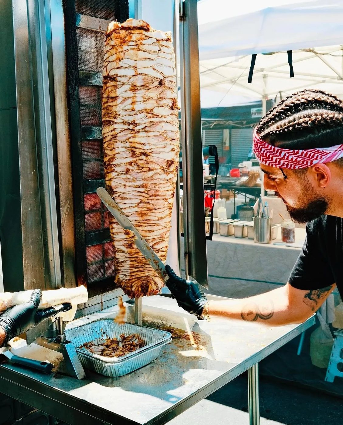 Cook in a red-and-white keffiyeh headband shaving a vertical shawarma spit into a foil tray at an outdoor market stall